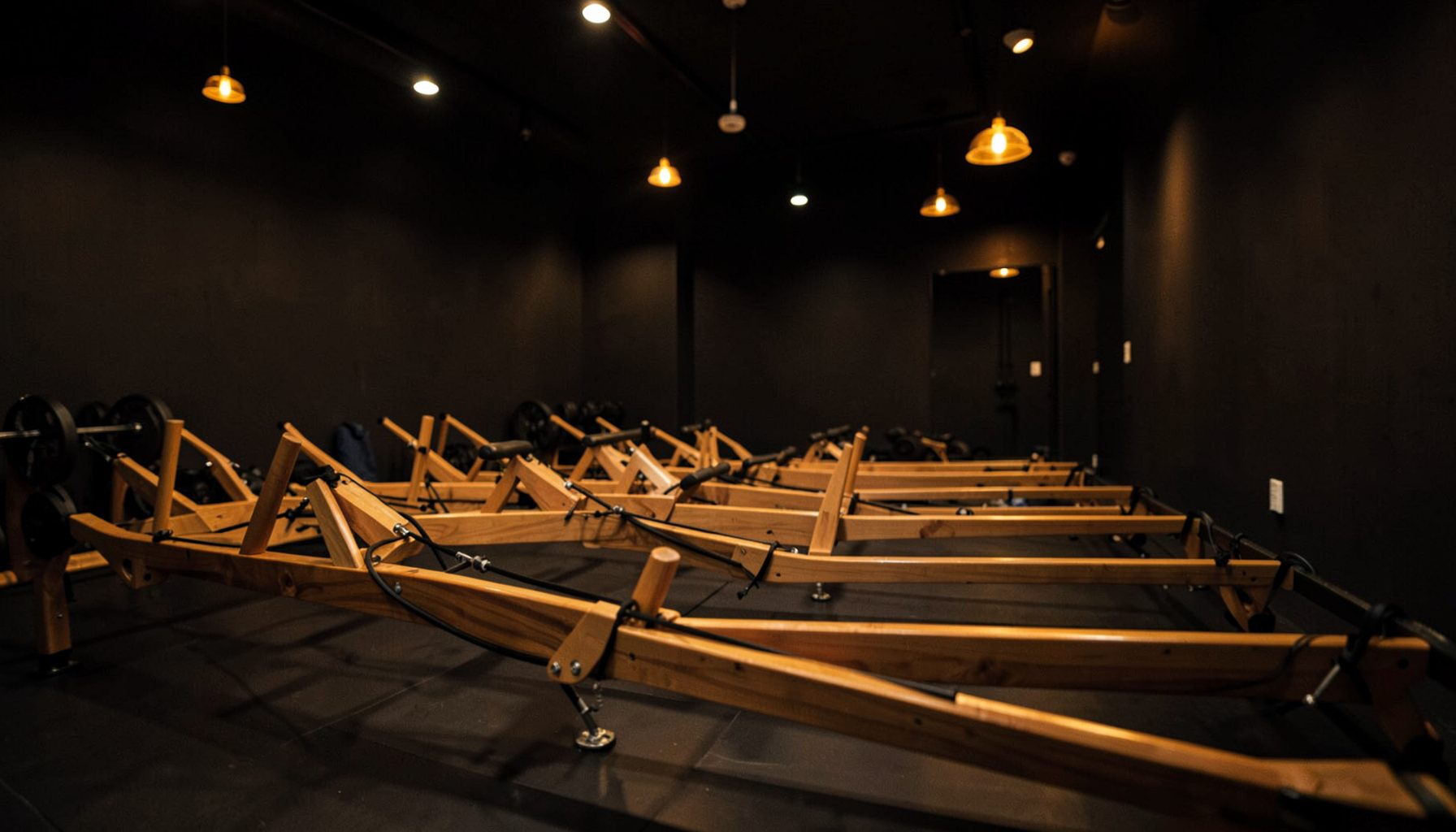 A dimly lit boutique fitness studio with rows of wooden water rowing machines, warm amber pendant lighting, and dark matte walls creating an intimate, high-energy atmosphere.